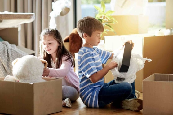 Young children opening cardboard boxes filled with toys and a tablet while sitting on the wooden floor in a cozy, sunlit home setting.