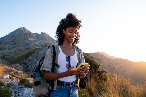 A young woman with a backpack using her smartphone during a hike in a mountainous area at sunset, illustrating the importance of travel health insurance for travelers abroad.