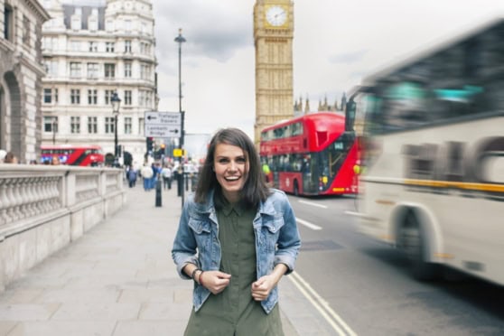 Young woman smiling in London street with Big Ben and double-decker buses, emphasizing international health coverage and travel safety for global citizens.