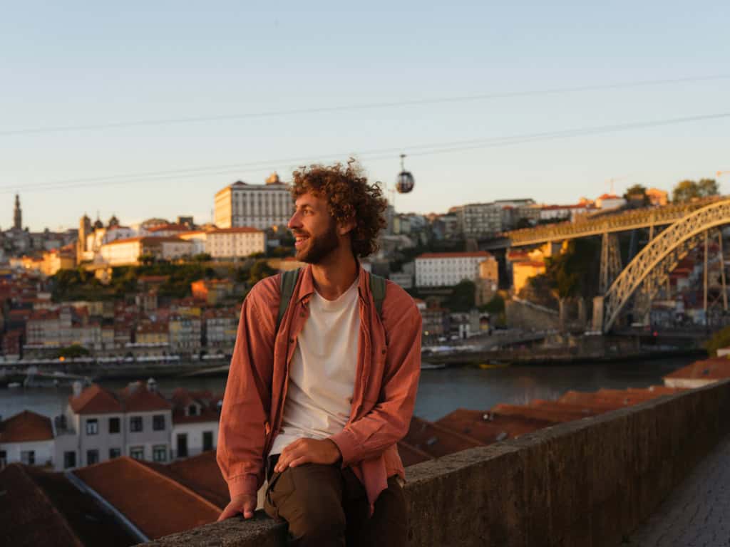 A young man with curly hair and a backpack relaxing on a ledge overlooking Lisbon, Portugal, during golden hour with iconic bridge, colorful rooftops, and city skyline.