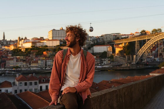 A young man with curly hair and a backpack relaxing on a ledge overlooking Lisbon, Portugal, during golden hour with iconic bridge, colorful rooftops, and city skyline.