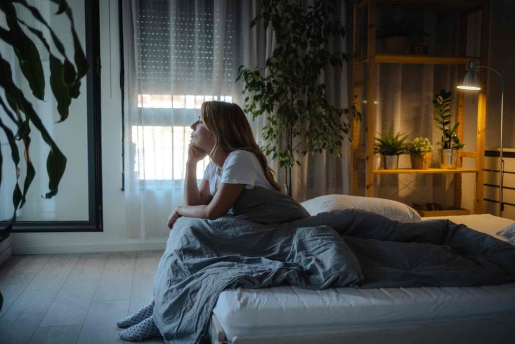 A woman sitting alone on her bed, looking out the window in a dimly lit bedroom, illustrating contemplation or concern, with indoor plants and warm lighting creating a cozy atmosphere.