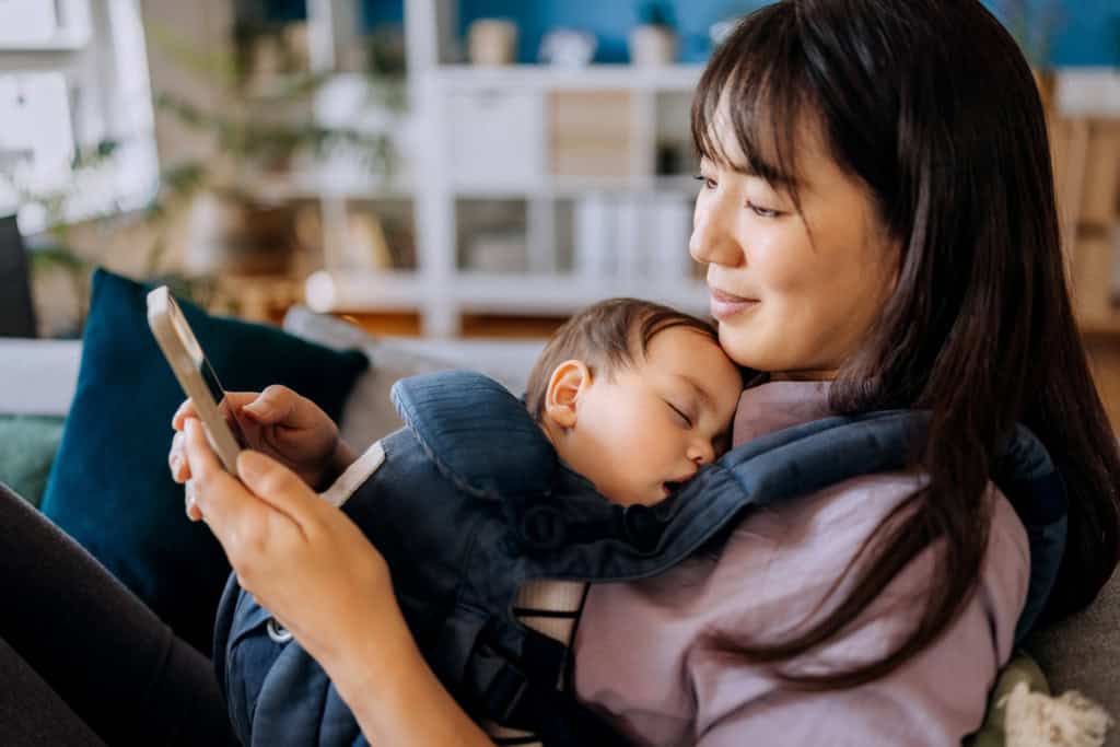 A mother relaxing with her sleeping baby in a baby carrier while using her smartphone in a cozy home setting. The background features blurred furniture and decor, emphasizing a peaceful moment of bonding and relaxation.