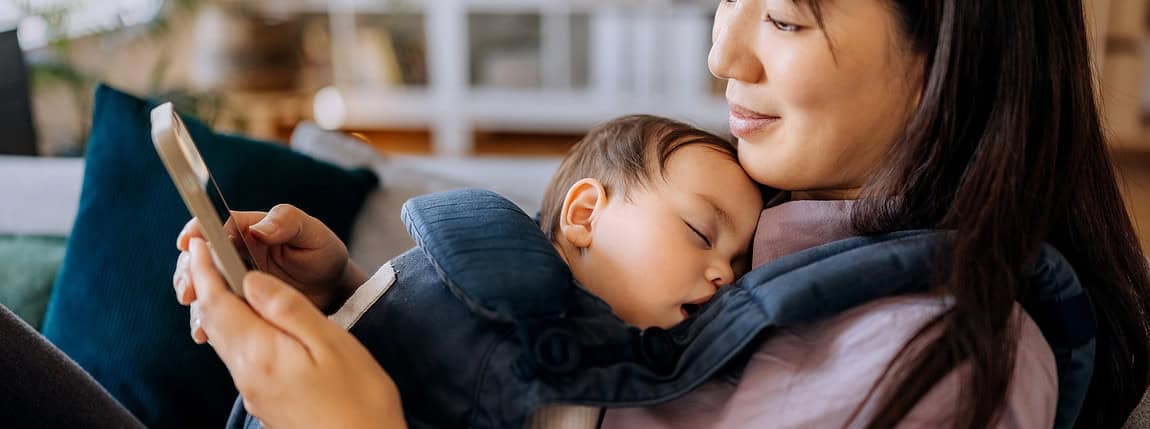 A mother relaxing with her sleeping baby in a baby carrier while using her smartphone in a cozy home setting. The background features blurred furniture and decor, emphasizing a peaceful moment of bonding and relaxation.
