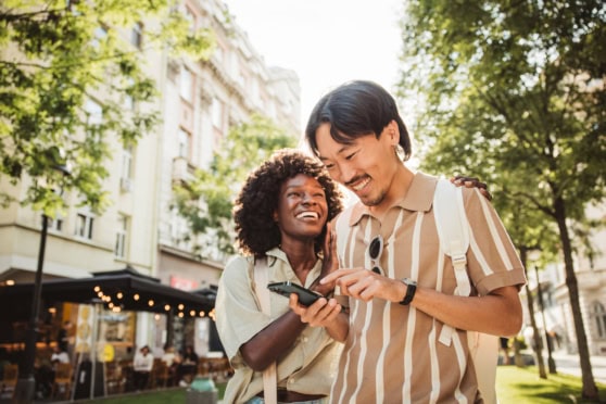 Smiling diverse travelers enjoying outdoor city life with mobile device, promoting global health coverage for international citizens.