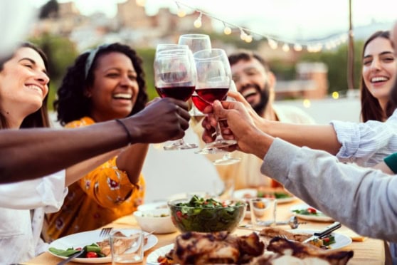 Friends of various backgrounds clinking glasses filled with red wine during a cheerful outdoor dinner on a cozy evening with string lights.