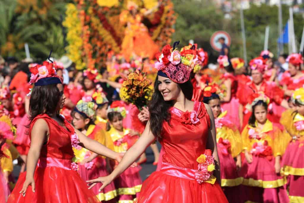 Brightly dressed women wearing flower-decorated hats and red dresses participate in a lively cultural festival, surrounded by children and onlookers.