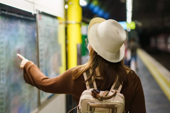 A woman with a hat and backpack points at departure information boards at an airport, illustrating international travel and insurance solutions for global citizens.