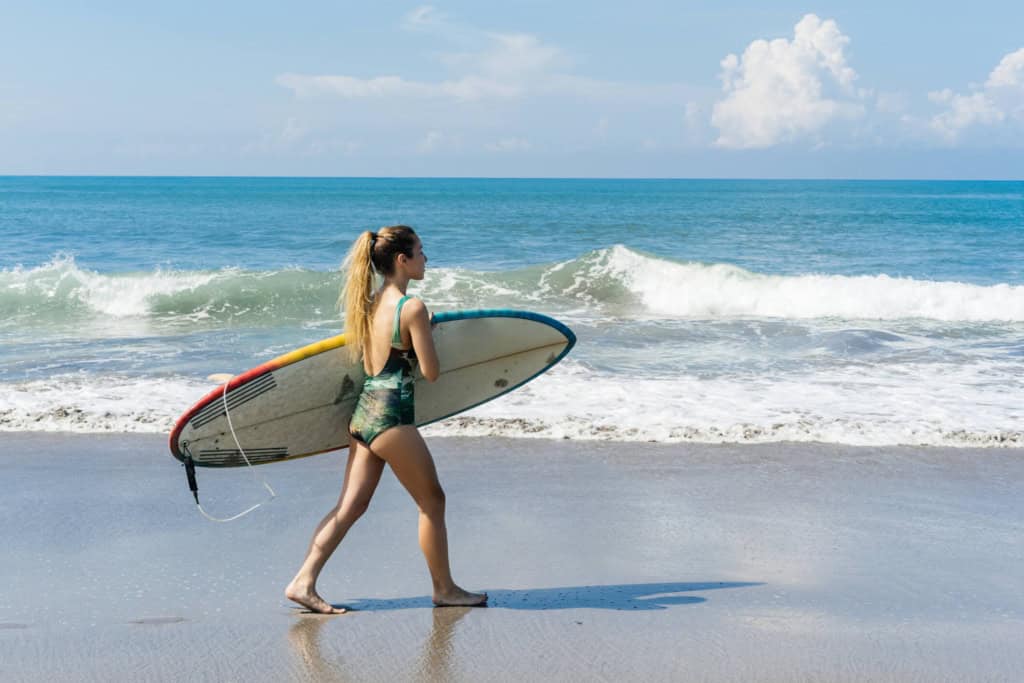 Young woman with a surfboard walking along the beach blue ocean waves and clear skies in background.