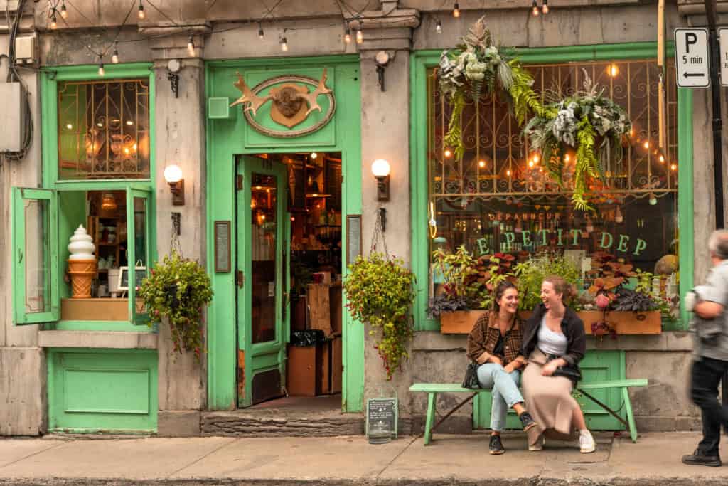 Street scene in Montreal, an LGBT friendly city in Canada. A charming café storefront with green-painted doors and window frames, decorated with hanging plants and string lights, creating a warm and inviting atmosphere.