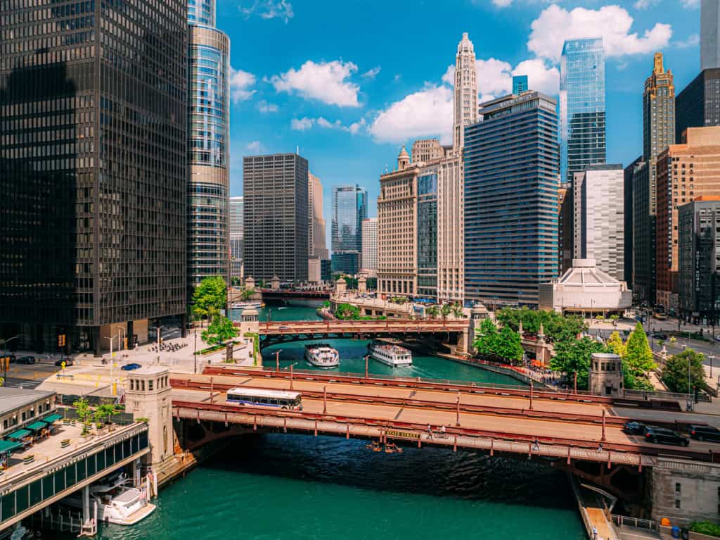 NYC skyline with tall buildings and river in the foreground, showcasing a vibrant urban landscape with bridges and boats under a bright blue sky.