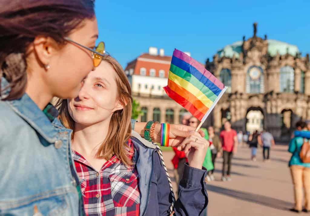 Two women at German Pride Parade, one holding a rainbow pride flag, celebrating LGBTQ+ rights and diversity in a vibrant city square with historic buildings in the background.