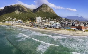 An aerial view of a vibrant beach with turquoise waters, sandy shores, and colorful umbrellas, set against the backdrop of lush green mountains and a lively coastal cityscape.