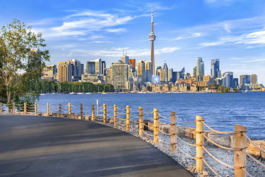 A vibrant cityscape of Toronto featuring the iconic CN Tower, modern skyscrapers, and a scenic waterfront promenade on a clear day.