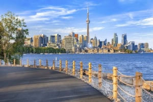A vibrant cityscape of Toronto featuring the iconic CN Tower, modern skyscrapers, and a scenic waterfront promenade on a clear day.