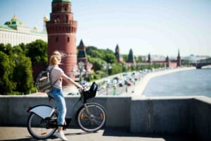 A woman with a backpack riding a bicycle along a bridge in Moscow, Russia, with the Kremlin and historic buildings in the background, showcasing urban travel and exploration.