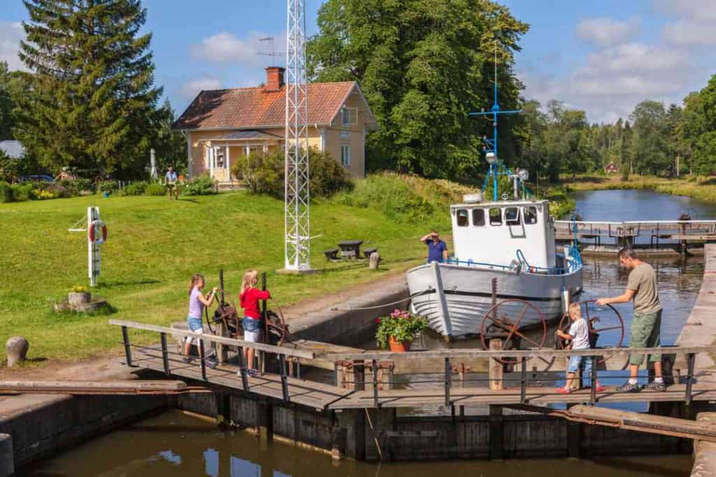 An image of a canal lock in Sweden with a small boat being guided through by people, surrounded by lush greenery and a charming house in the background.