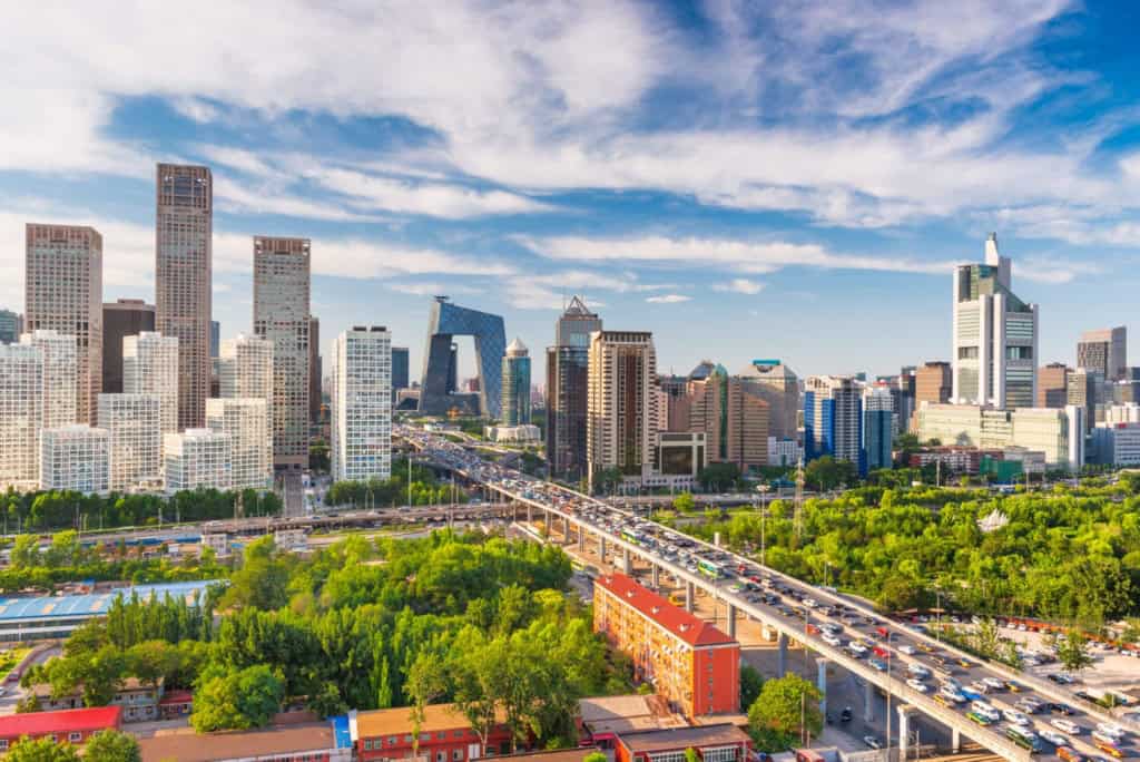 Urban cityscape featuring tall buildings, a busy highway, and lush green areas under a bright blue sky.