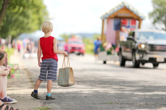 Child holding a shopping bag watching a small-town parade with floats and families on a sunny day.