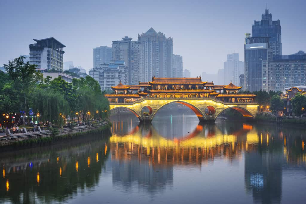 An image of a traditional Chinese bridge illuminated at night, reflecting on the river, with modern skyscrapers in the background, representing the blend of history and modernity in China.