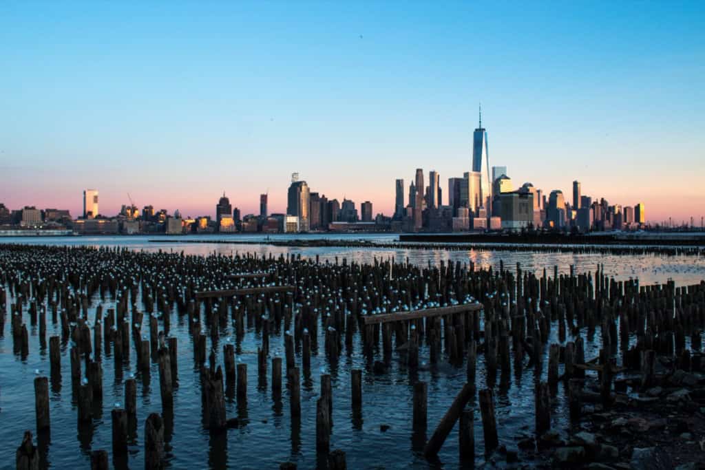 Iconic New York City skyline with One World Trade Center at sunset, viewed from the waterfront with wooden pilings in the foreground.