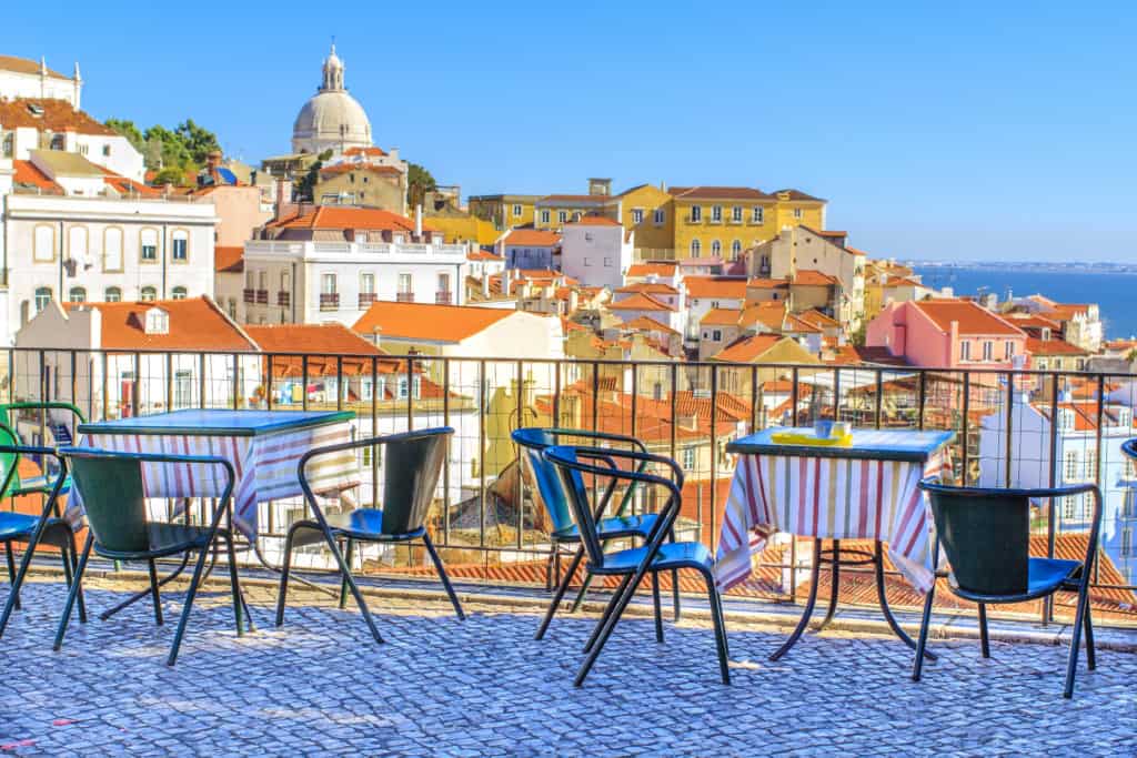 Scenic terrace in Lisbon with colorful tables and chairs, offering a panoramic view of the city's rooftops, historic buildings, and the distant river under a clear blue sky.