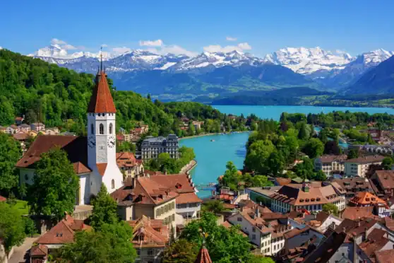 An aerial view of a picturesque Swiss village featuring a historic church, colorful rooftops, lush greenery, and a winding river against a backdrop of snow-capped mountains.