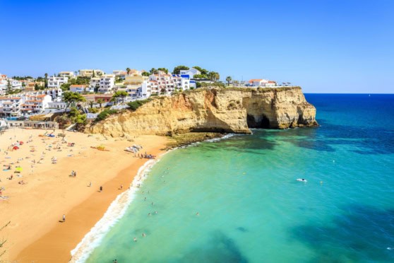 Scenic view of golden cliffs and turquoise waters at a beach in Algarve, Portugal, with white buildings on the cliffs and a clear blue sky.