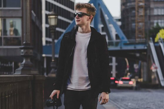 Young man with sunglasses walking in city, holding camera, representing US citizens preparing for international travel.