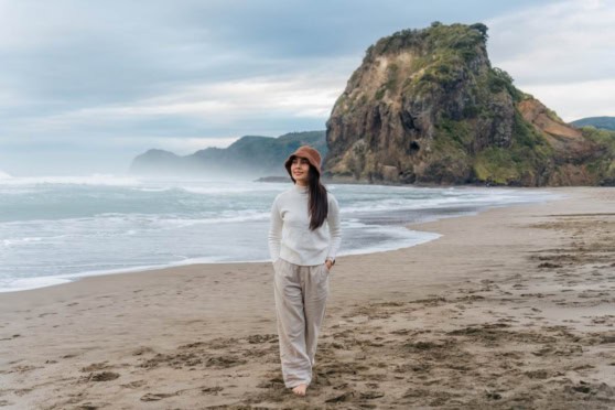 A woman standing on a sandy beach with a large rocky cliff in the background, under a cloudy sky, representing travel and international exploration.