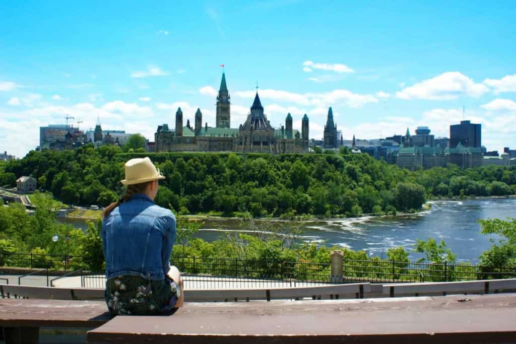 An individual sitting on a bench overlooking the city skyline of Ottawa, Canada, with Parliament Hill and the Rideau River visible under a bright blue sky.