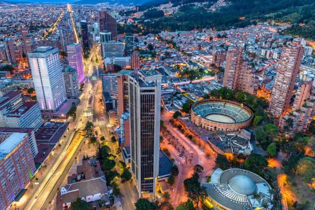 A vibrant aerial view of Bogotá, Colombia, showcasing tall skyscrapers, busy streets, and the historic Monserrate hill illuminated at sunset.