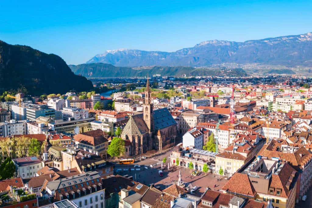 An aerial view of Bolzano, Italy, showcasing its historic city center, colorful buildings, and surrounding mountains, highlighting the city's safety and scenic beauty.