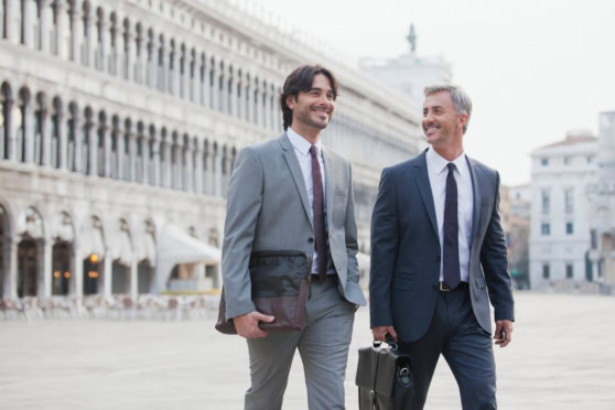 Two professional men in business attire walking in an Italian city square, discussing work visas and international insurance options for Italy.
