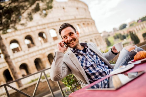 Happy American man enjoying a phone call at a cafe near the Colosseum in Rome, Italy, highlighting work opportunities for Americans in Italy.