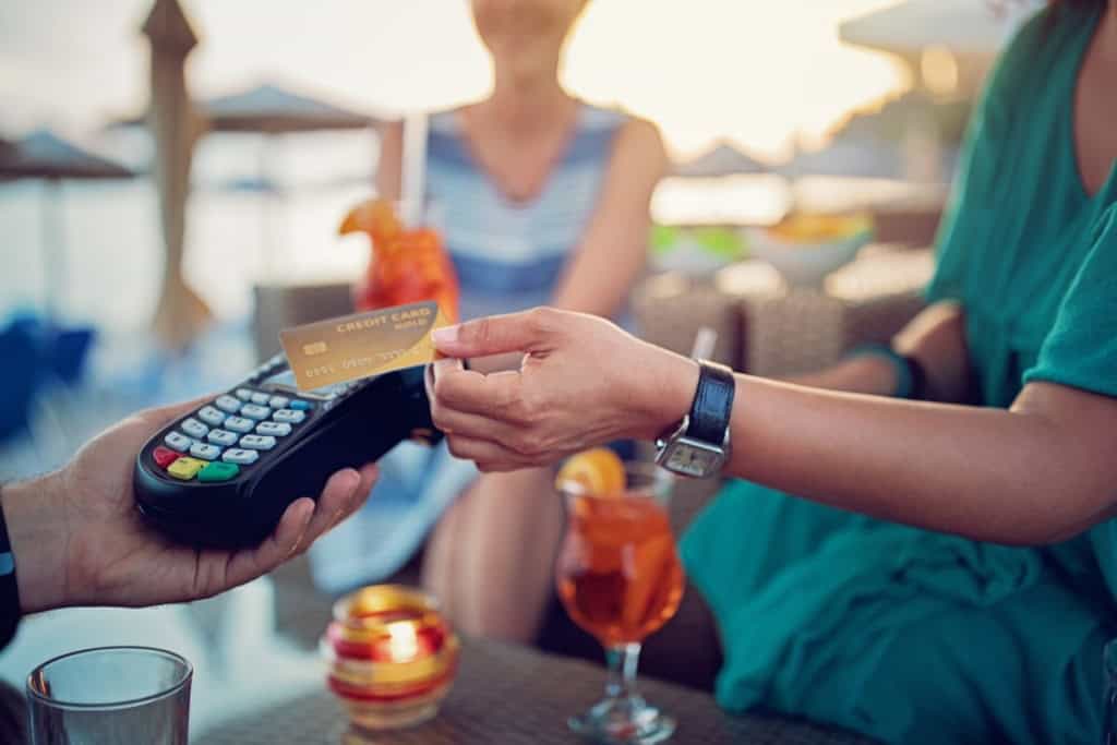 A person making a contactless payment with a credit card at an outdoor café in Greece, with a woman in the background.