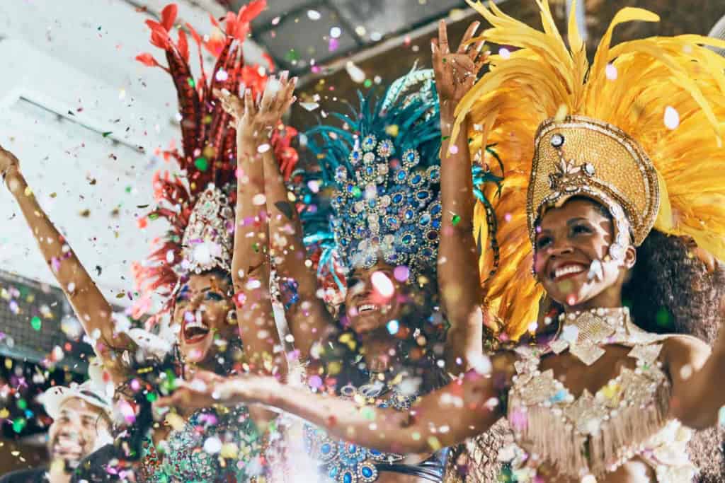 An energetic group of carnival dancers in colorful costumes with feathers and sequins, celebrating joyfully with confetti and bright smiles.