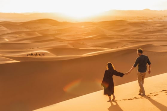 An adventurous couple holding hands while walking across golden sand dunes at sunset.