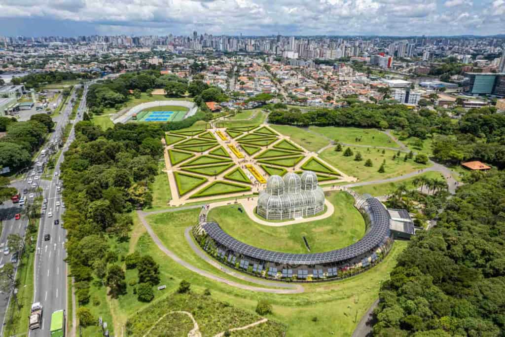 Aerial view of São Paulo's lush botanical gardens featuring geometric flower beds, glasshouses, and surrounding greenery, with the city skyline in the background.