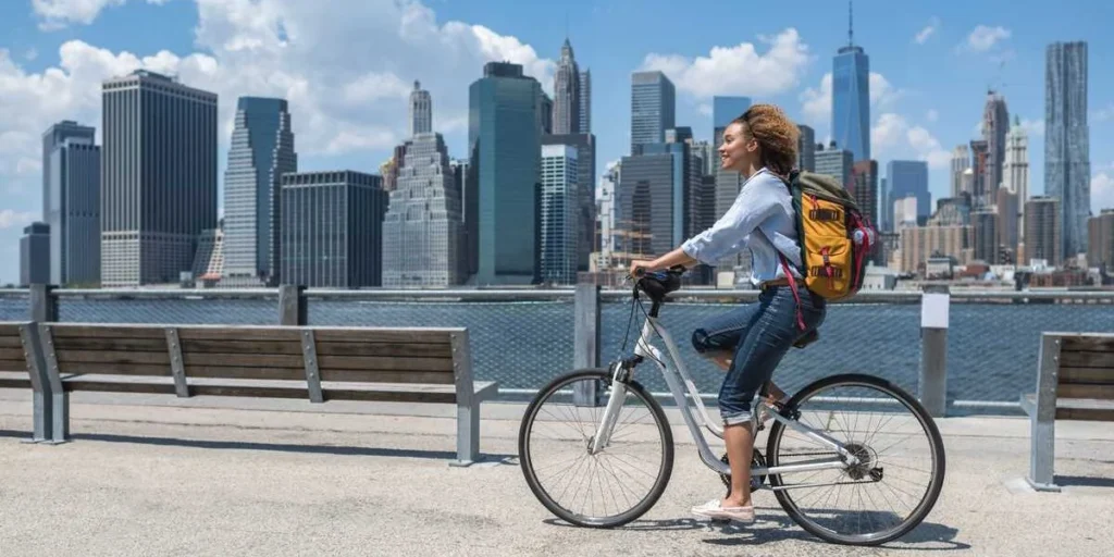 Young woman riding a bicycle along Manhattan waterfront with New York City skyline in background, representing international residents in the U.S.