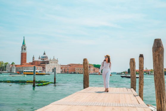 A woman in white standing on a wooden dock in Venice, Italy, with historic buildings and the lagoon in the background.