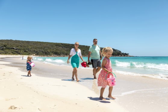 Family enjoying a sunny day at the beach in Australia, with children playing in the sand and adults walking along the shoreline under a clear blue sky.