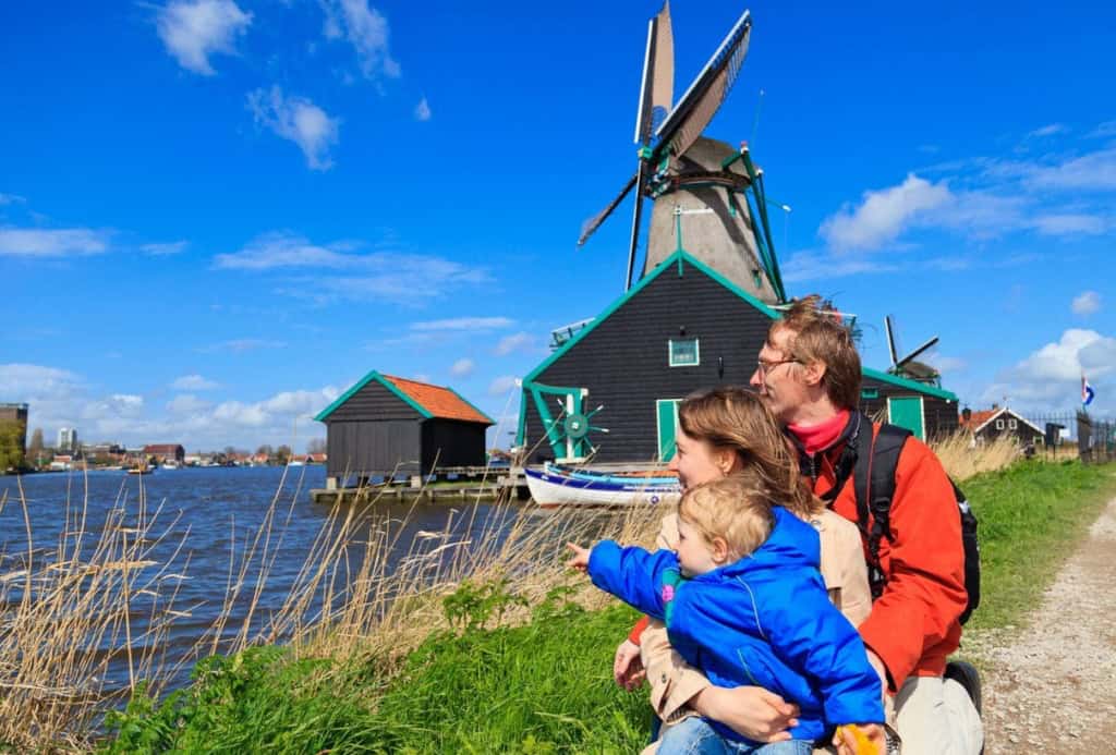 A family enjoying a scenic view of a traditional Dutch windmill near a canal in the Netherlands, with clear blue skies and lush greenery, highlighting the country's iconic landscape.