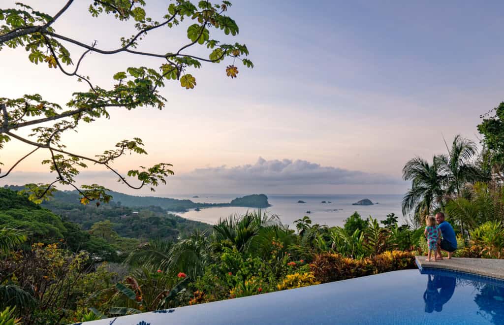 A scenic view of Costa Rica's lush tropical landscape with a distant ocean horizon, surrounded by vibrant greenery and palm trees, under a partly cloudy sky.