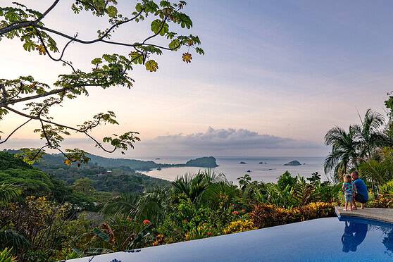 A scenic view of Costa Rica's lush tropical landscape with a distant ocean horizon, surrounded by vibrant greenery and palm trees, under a partly cloudy sky.