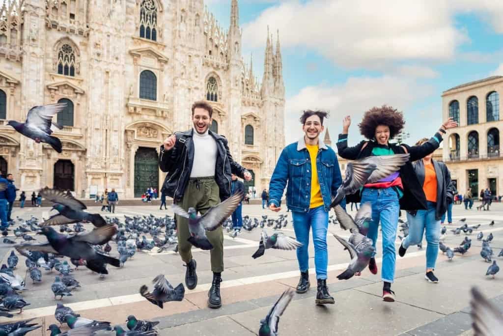 Group of diverse young adults enjoying and feeding pigeons in Piazza del Duomo, Milan, during daytime.