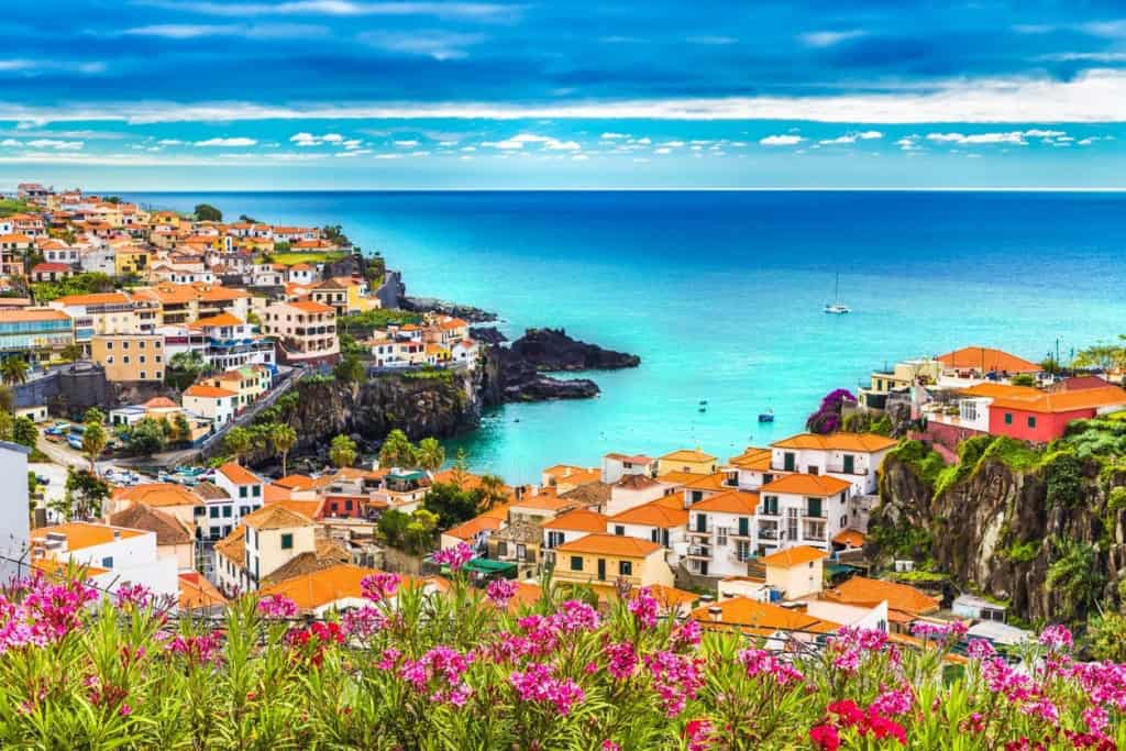 Scenic view of a colorful coastal village with vibrant houses, lush greenery, and a sparkling blue sea under a partly cloudy sky, perfect for expatriates seeking a Mediterranean lifestyle.