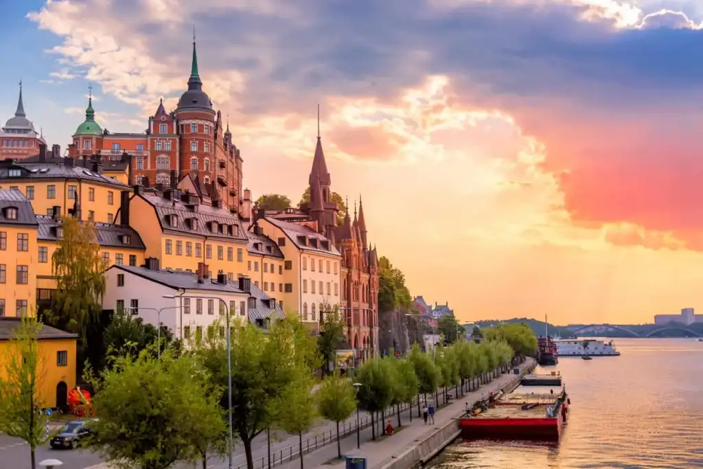 An image of a picturesque Swedish cityscape along a waterfront, featuring colorful historic buildings, lush greenery, and a serene river at sunset, highlighting the charm of living abroad.