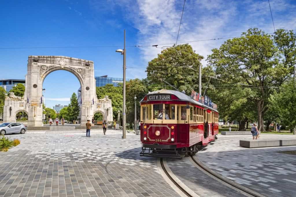 An image of a vintage red tram traveling through a city square with historic archway and lush green trees, representing international living and travel opportunities.