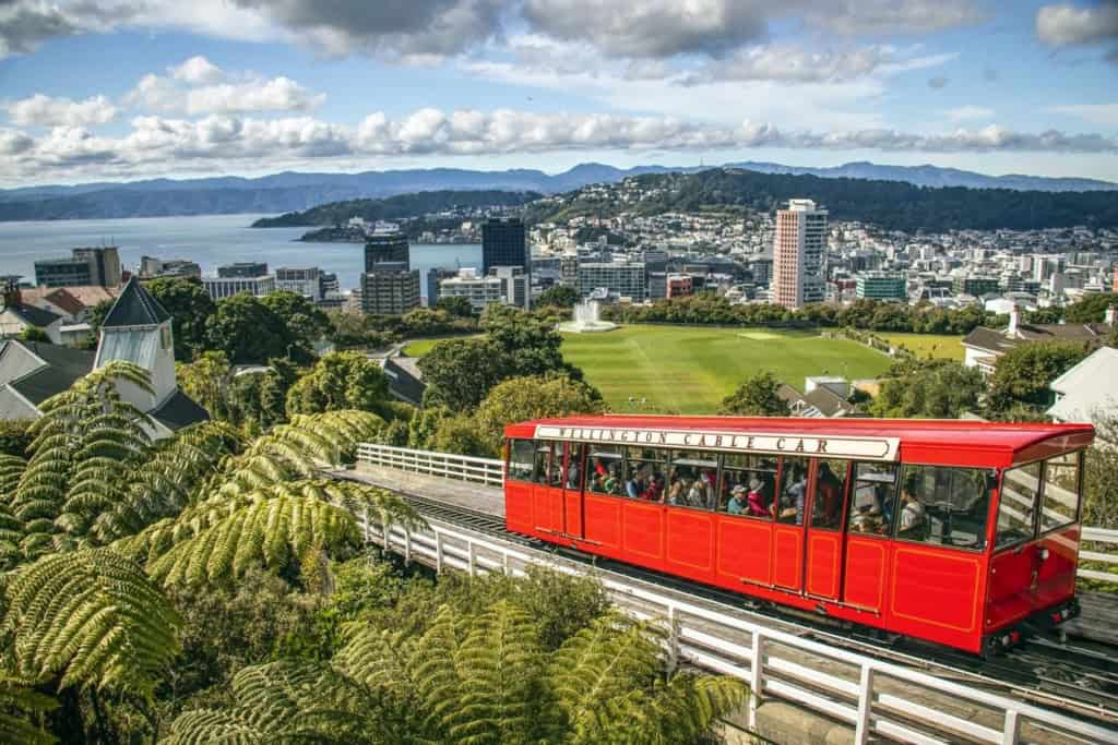 A red cable car ascending through lush greenery with a panoramic view of Wellington, New Zealand, featuring city buildings, green parks, and distant mountains under a partly cloudy sky.